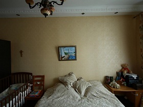 мягкие игрушки на столе с полкой и шкафчиком, большая кровать, стул и детская кроватка у бежевой стены спальни квартиры времен СССР