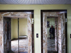 лифтовые кабины с зеркальными дверцами, обрамленные мраморной плиткой в престижном подъезде современной многоэтажки