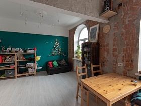 деревянный обеденный стол у кирпичной стены с деревянными часами, колонкой на деревянной полке в просторной кухне зонированой комнаты со светло серым полом