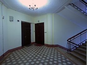 небольшие коврики у дверей соседних квартир на полу с бежево коричневой фигурной плиткой, белыми стенами и большой люстрой на потолке