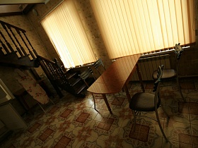 коричневый обеденный стол со стульями у окон с закрытыми вертикальными белыми жалюзи на окнах кухни двухэтажного дома под съем в сосновом лесу