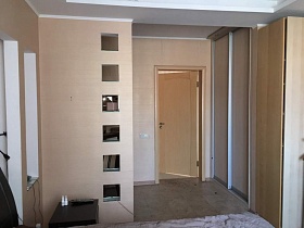 прикроватная коричневая темная тумбочка у разделительной стены из гипсокартона с открытыми квадратными окошками в спальной комнате семейной квартиры