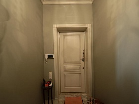 круглый коричневый небольшой столик на фигурных ножках у стены с видеодомофоном, коврик на полу у светлой входной двери бежевой прихожей современной дизайнерской квартиры