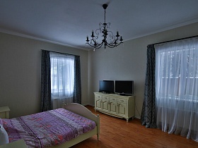 два телевизора на белой тумбочке, белая большая кровать с цветным покрывалом в светлой спальне с темно синими шторами современного кирпичного дома
