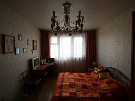 общий вид спальной комнаты с компьютерным столом у большого окна с белой гардиной и сиреневыми шторами, ярким оранжево-сиреневым покрывалом на большой кровати, картинами на светлых стенах просторной трехкомнатной квартиры