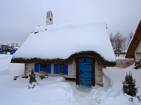 расчищенная тропинка к домику мазанка с синими дверьми и ставнями на окнах и небольшими елочками в сугробах снега на территории ресторана, стилизованного под хутор в коттеджном поселке