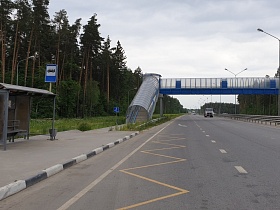 широкая автомобильная трасса с крытой остановкой и голубым подвесным мостом с арочным прозрачными перекрытием над дорогой в лесу для съемок кино