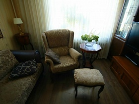 ваза с цветами на деревянном восьмигранном столике на фигурных ножках, мягкий диван, кресло с подушками и пуфик в бежевом цвете у окна с гардиной гостиной квартиры оператора