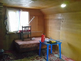канистра на синем пластиковом столике, кровать с матрасом у окна, кирпичная труба печи в комнате с цветным ковром на полу деревянного домика в деревне