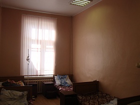 белые вертикальные жалюзи на высоком окне палаты с персиковыми стенами, освещением на белом потолке, деревянными кроватями на полу с коричневым линолеумом действующей старой больницы