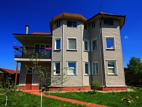общий вид семейного дачного яркого трехэтажного дома на зеленом участке за высоким забором с видом на город