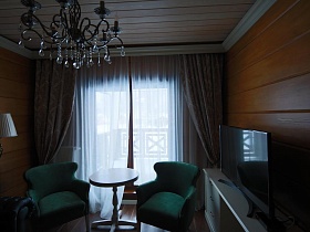 круглый столик на ножке и два зеленых кресла у окна в гостиной деревянного съемного коттеджа