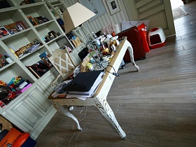 заставленный разнообразными предметами журнальный столик на фигурных резных ножках, стул - кресло у белого открытого книжного шкафа в гостиной дизайнерской квартиры с видом на Москву реку