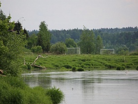 белые футбольные ворота на зеленой поляне на берегу реки со спокойной поверхностью воды в окружении зелени