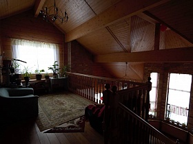 за деревянными перилами небольшая комната для отдыха с диванчиком, креслом и комнатными цветами у окна