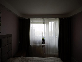 белая гардина и коричневые шторы на окне с балконной дверью спальной комнаты современной трехкомнатной квартиры в светлых тонах