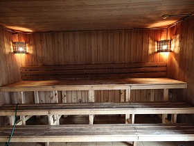 светильники с деревянными решетками в углах парной с деревянными полками в три яруса в бревенчатом доме банного комплекса