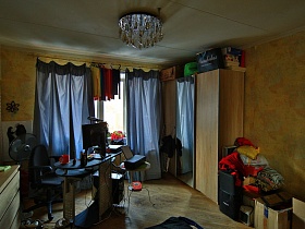 многочисленные коробки и ящики на полу, светлый шкаф для одежды с зеркалом посередине у окна с серыми шторами, черное кресло у компьютерного стола, вентилятор в гостиной с круглой под потолок люстрой