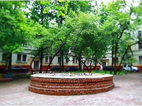 круглый пустой фонтан на просторной площадке с зелеными деревьями по периметру в старом дворе трехэтажного дома