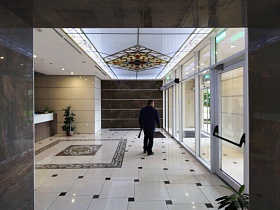 красивый престижный подъезд элитной многоэтажки с белой квадратной плиткой на полу, натяжным потолком, комнатным цветком у ресепшн, большими стеклянными дверьми
