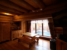 самовар и настольная лампа на столешнице деревянной кухонной мебели, скамейки вокруг прямоугольного стола в зоне чайной с большими окнами домика из сруба
