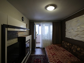 двухцветная мебельная стенка напротив разложенного дивана с коричневым покрывалом, круглым закрытым плафоном подвесной люстры на белом потолке спальной комнаты с двухцветными стенами