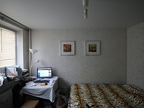 картины под стеклом в рамках на стене, белый торшер в стиле хай-тек,стол с монитором у окна с многочисленными бумагами на подоконнике светлой спальни современной двухкомнатной квартиры