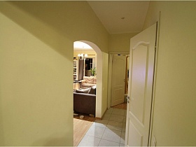 коричневый и бежевый мягкие диваны с подушками через арочный дверной проем светлого коридора белой трехкомнатной квартиры