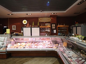 мясная продукция и колбасные изделия под стеклом больших витринных холодильников у коричневой стены с высокими шкафами в здании продуктового районного магазина