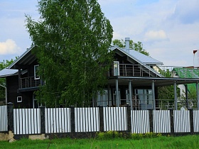 открытый просторный балкон над большой террасой с колоннами современного элитного дома темно серого цвета с белой крышей за белым забором в живописном месте Подмосковья