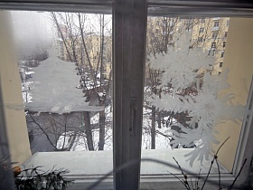 вид на ветви высоких деревьев на заснеженном дворе через стекла окна с наклееными белыми снежинками трехкомнатной сталинской квартиры эпохи СССР