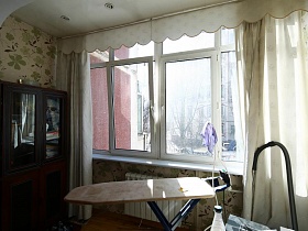 гладильная доска, темный шкаф  у большого окна с белыми шторами застекленной лоджии простой семейной квартиры в Марьино