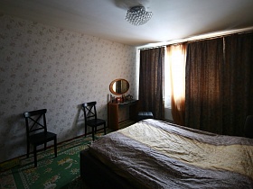 просторная светлая спальная комната с темной мягкой кроватью, деревянными стульями у стены, зеркалом над гримерным столиком у окна с коричневыми шторами и гардиной современной квартиры
