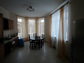 круглый обеденый коричневый стол со стульями, мебельная стенка в просторной кухне с высокими окнами кирпичного двухэтажного дома под съем