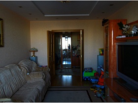 серый телевизор в рыжей стенке,картина над белым диваном и детские машинки у входной двери в гостиную комнату приличной трешки панельного дома