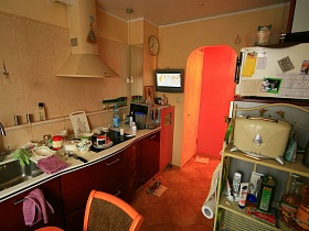 вишневая кухня с белой рабочей поверхностью, этажерка с кухоннми предметами ,телевизор и часы у арочного дверного проема в семейной трешке