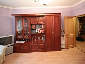 открытая дверь в сиреневую гостиную с коричневой стенкой, серым телевизором на сером комоде и бежевой мягкой мебелью квартиры молодоженов в современном доме