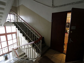 квадратная плитка на полу лестничной площадки перед открытой дверью в жилую квартиру в подъезде дома с большим окном и ступенями с перилами между этажами