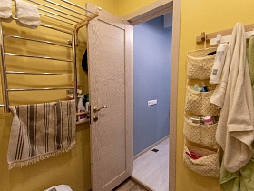 полосатое полотенце на полотенцесушителе, подвесной модульный органайзер и полотенца на настенной деревянной вешалке на желтых стенах ванной комнаты у открытой двери современной квартиры