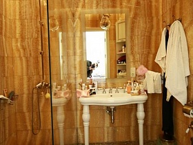 белая раковина на фигурных ножках с туалетными принадлежностями и прямоуголным зеркалом на стене, санузел, одеждв на настенной вешалке, душ за стеклянной стеной в ванной комнате с коричневой мозаичной плиткой на полу девчачьей двухкомнатной квартиры