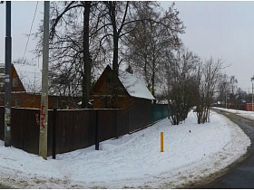 дорога вдоль двух желтых угловых дома с коричневым высоким забором