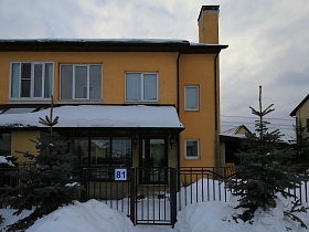 небольшие елочки под снегом у металлического забора с номером у ворот вокруг двухэтажного желтого дома