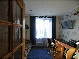 кресло и компьютерный стол с принтером у светлой стены с рисунком,синий коврик у дивана, синие шторы на окне светлой комнаты евро квартиры с видом на Москву и парк