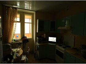 белый холодильник, круглый обеденный стол и салатовая мебельная стенка в кухне с желтыми стенами и желтой гардиной на окне с балконной дверью современной двухкомнатной квартиры