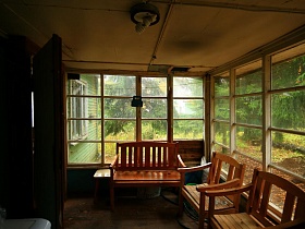 деревянный диван, кресла и табурет у окон веранды жилой лачуги среди высоких елей у пруда