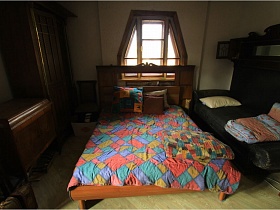 деревянный комод, шкаф, большая кровать с цветным покрывалом у необычной формы окна сказочного дома в лесу