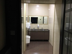 белое ведро для мусора, электросушилка для рук на стене с бежевой плиткой, большое прямоугольное зеркало над шкафом с мойкой и кранами в туалетной комнате ресторана