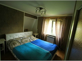 большая кровать с белой мягкой спинкой и голубыми покрывалами на цветном постельном, белые высокие прикроватные тумбочки в спальне с темными стенами стильной дачи для съемок кино