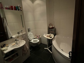 серый комод, треугольный столик у белой угловой ванны, большое зеркало над белой раковиной, санузел на сером полу ванной комнаты с белой плиткой на стенах