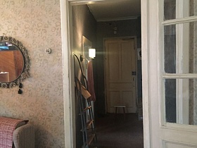 круглое зеркало в ажурной рамке на стене над мягким креслом прихожей с бежевыми обоями и открытой дверью в коридор с лестницей стремянкой, табуретом и бра на стене